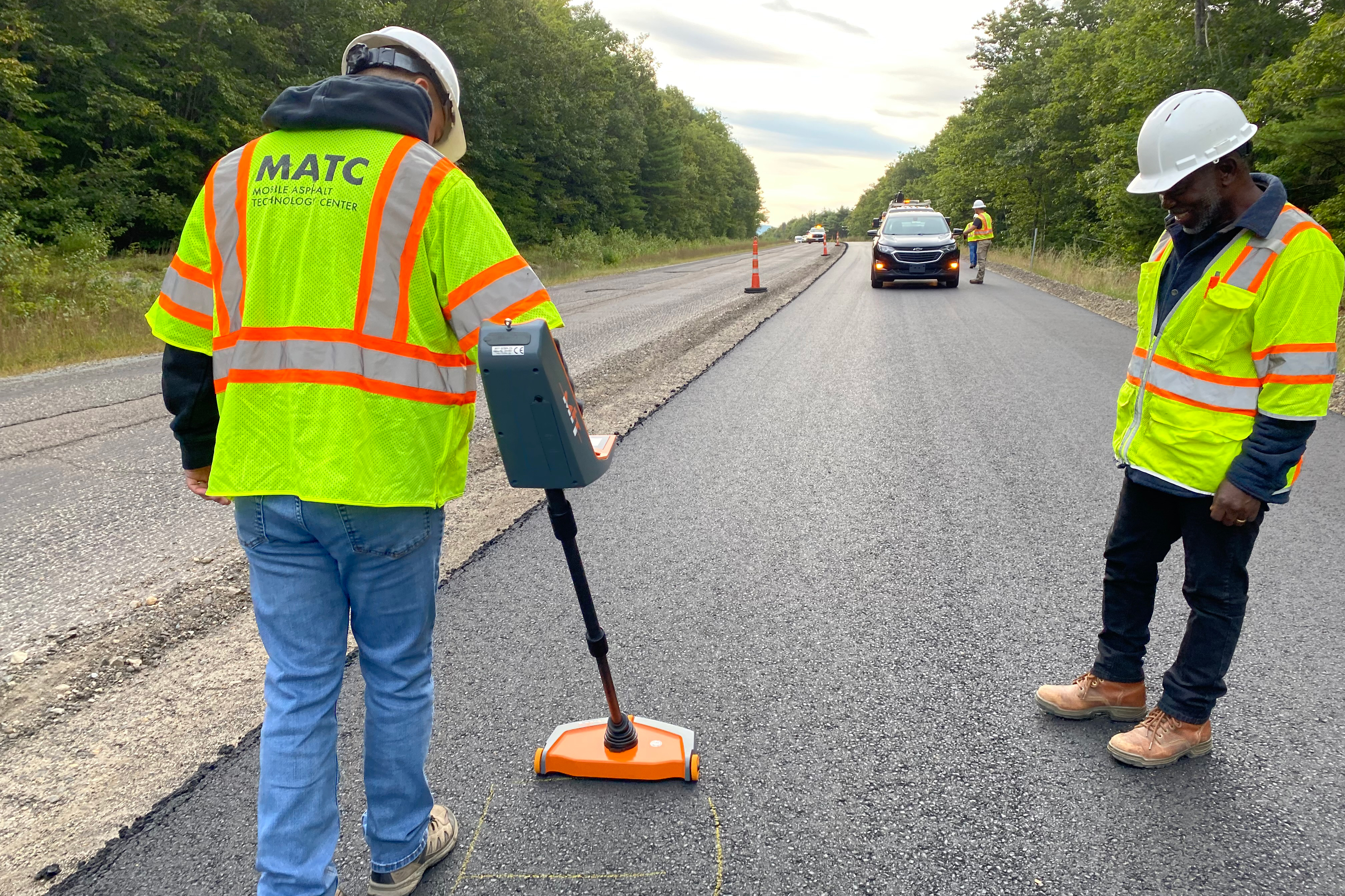 Two men in construction vests on an active asphalt paving process who are running a pulse induction test